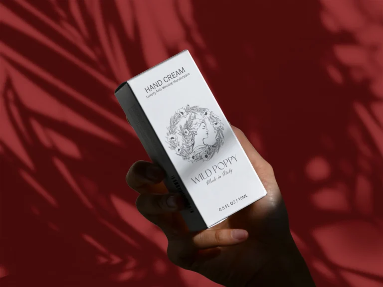 O mână care ține o cutie albă de ambalaj pentru crema de mâini Wild Poppy, imprimată cu o emblemă ilustrată în detaliu, pe un fundal roșu burgund cu umbre de frunze, realizată de FloBold.