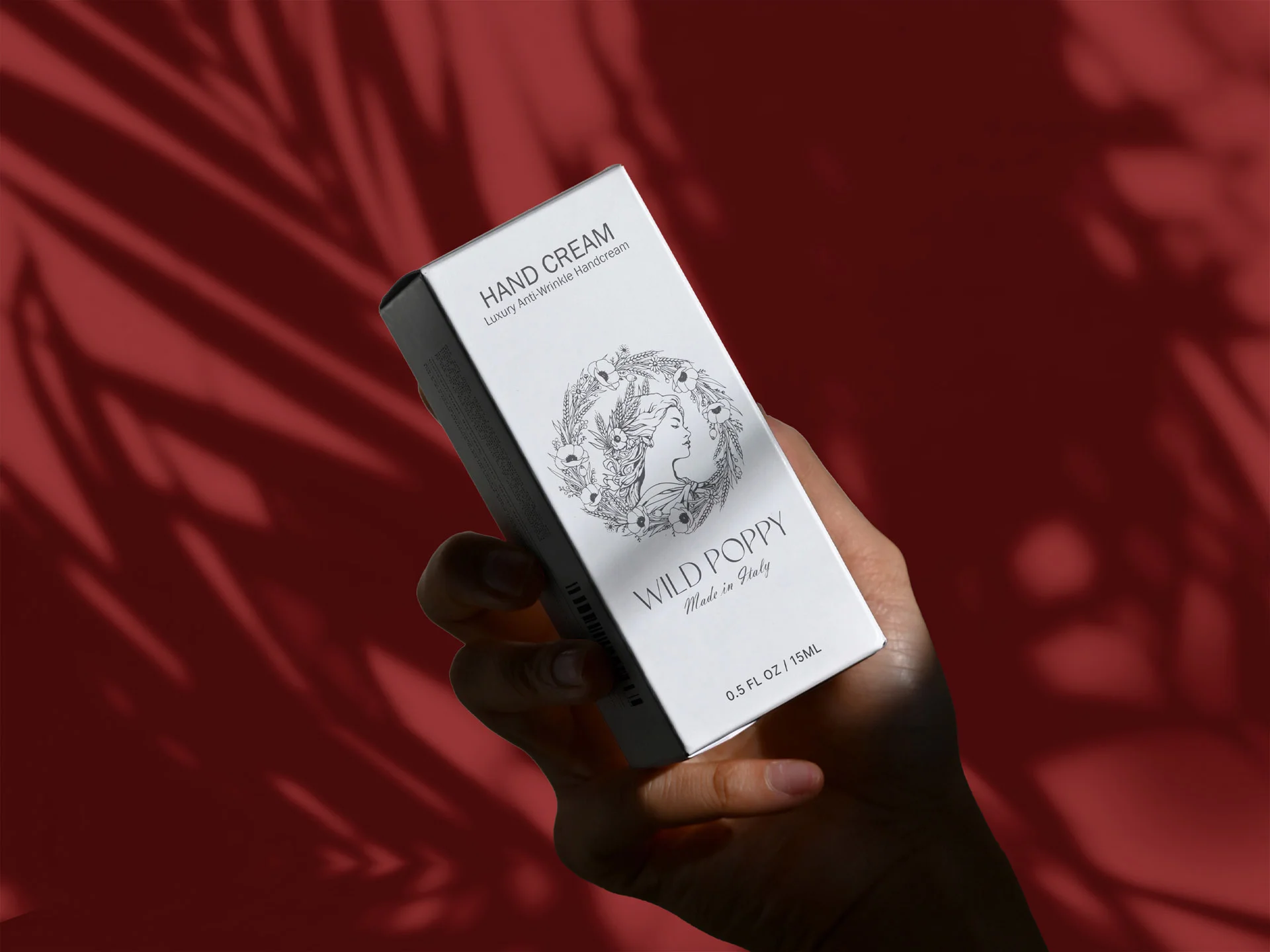O mână care ține o cutie albă de ambalaj pentru crema de mâini Wild Poppy, imprimată cu o emblemă ilustrată în detaliu, pe un fundal roșu burgund cu umbre de frunze, realizată de FloBold.